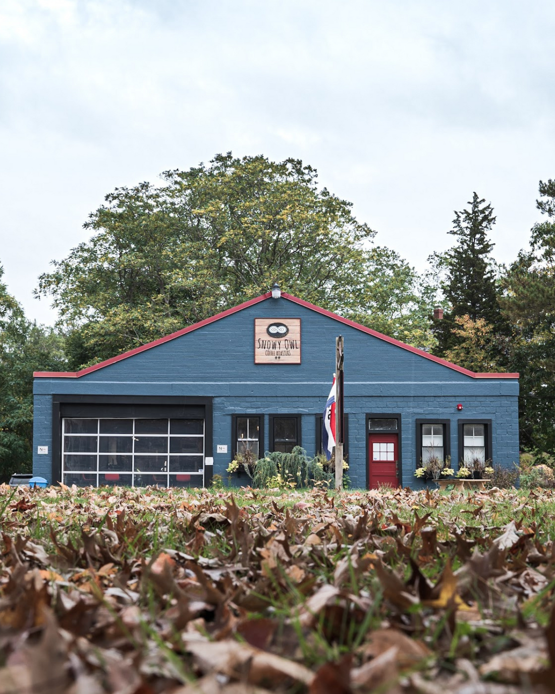 Snowy Owl Coffee Roasters' charming blue Sandwich cafe and roastery on Cape Cod, featuring cozy fall atmosphere with autumn leaves and signature red trim, serving fresh specialty coffee.