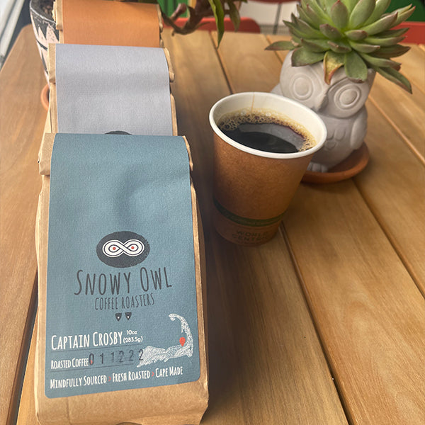 A freshly brewed cup of artisanal coffee sits alongside Snowy Owl's signature Captain Crosby blend, showcasing our mindfully sourced and locally roasted Cape Cod coffee beans.