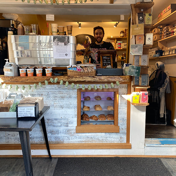 Inside Snowy Owl Coffee Roasters Chatham location with a cozy display of branded merchandise, coffee cups, and self-serve condiments, all arranged near a rustic counter and a window view of trees.